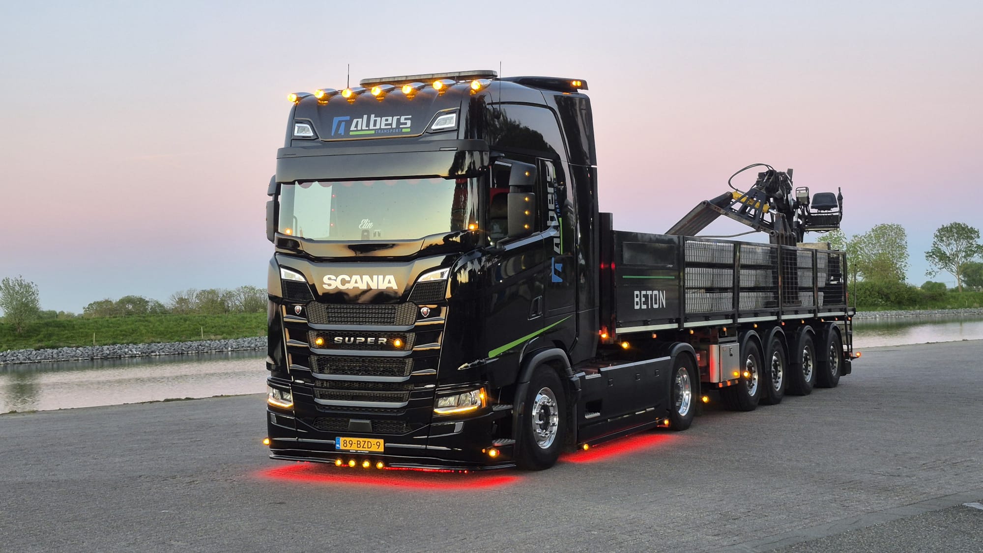 Zwarte Scania van Albers Transport met verlichting bij zonsondergang langs het water